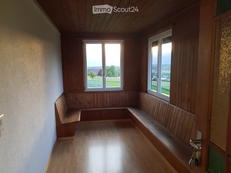 Empty room with wooden walls, wooden floor, built-in wooden benches along the walls, and large windows offering a view of the countryside.