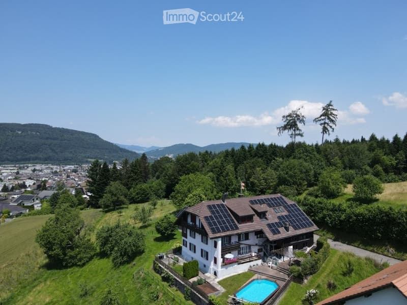 A two-story house with solar panels on the roof, surrounded by a well-maintained garden and a swimming pool. The house is situated on a hillside with a scenic view of the valley and mountains in the background.