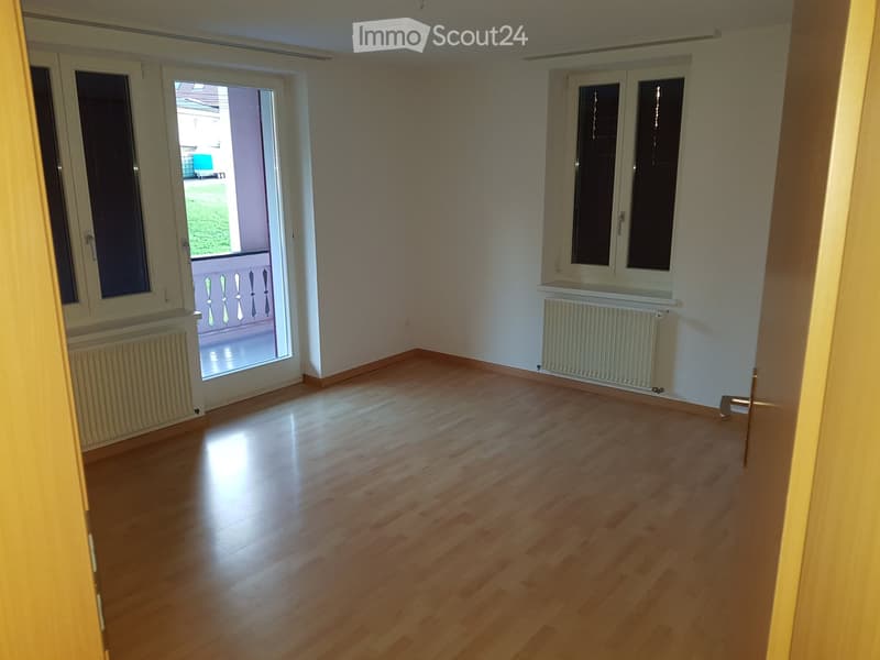 Empty room with wooden flooring, white walls, and large windows, including a sliding door leading to a balcony.