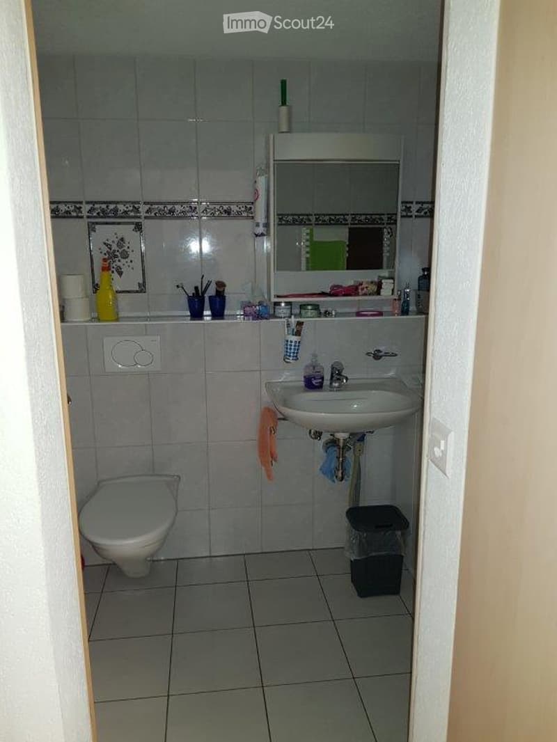 A small bathroom with a toilet, sink, mirror, and various toiletries on the counter. The walls are tiled, and the floor is also tiled.