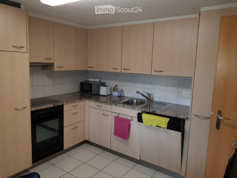 A compact kitchen with light wood cabinets, granite countertops, and tiled flooring. Visible appliances include a microwave, oven, and toaster. A sink with a faucet is present, along with a few towels hanging on the cabinet doors.