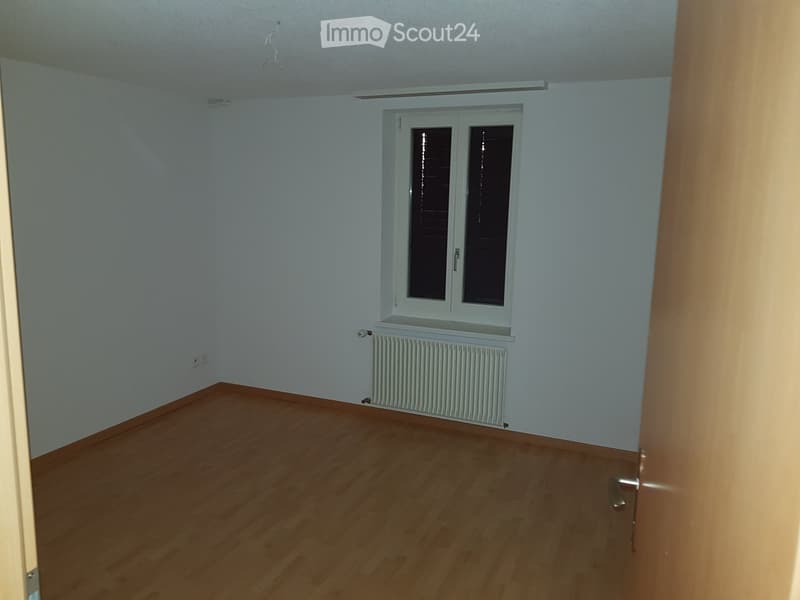 Empty room with light blue walls, wooden flooring, a window with blinds, and a radiator below it.