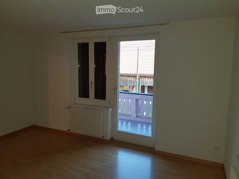 Empty room with wooden flooring, white walls, a radiator, and a large window with a view to a balcony.