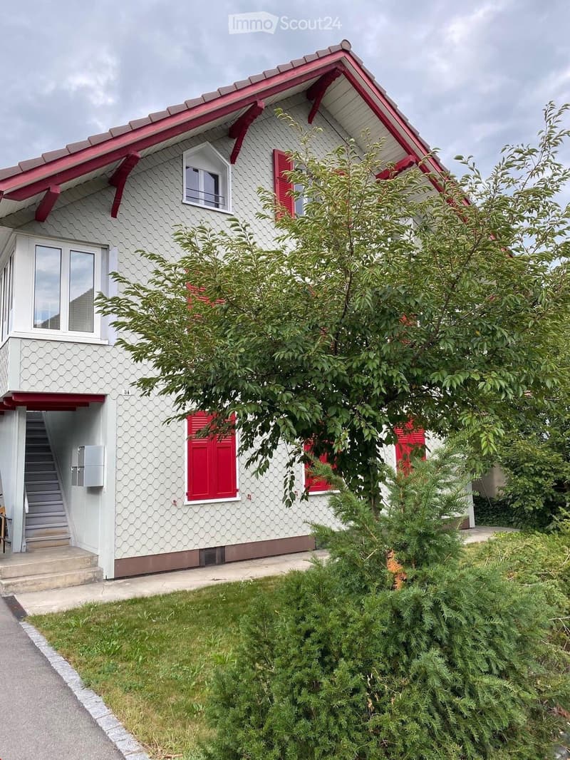 Two-story house with white siding, red shutters, and a red roof. Features a staircase leading to the entrance, surrounded by greenery and a well-maintained lawn.