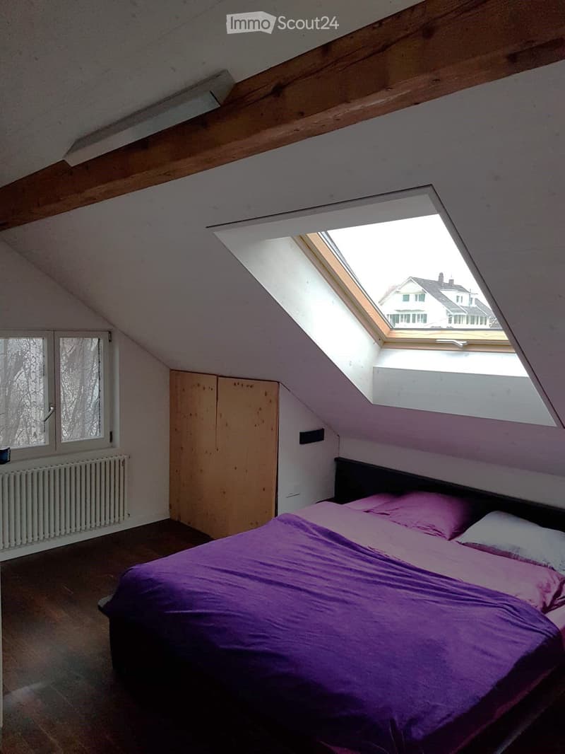 Bedroom with slanted wooden ceiling, skylight window, bed with purple bedding, wardrobe, radiator, and window with frosted glass.