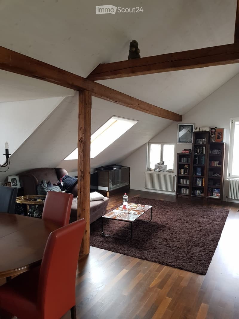 A cozy attic living space with wooden beams, skylights, and large windows. Features a brown sofa, coffee table, dining table with chairs, bookshelf, and a large area rug.