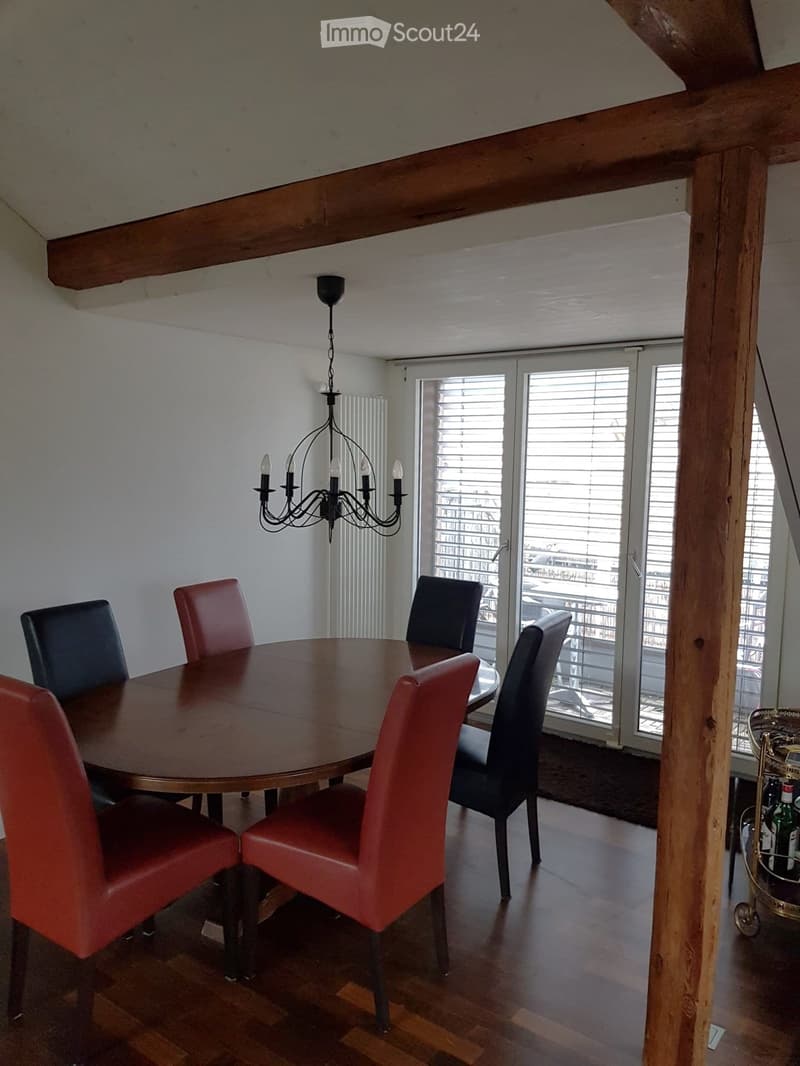 Dining room with a round wooden table surrounded by red and black chairs, a chandelier hanging from the ceiling, and large windows with blinds leading to a balcony.