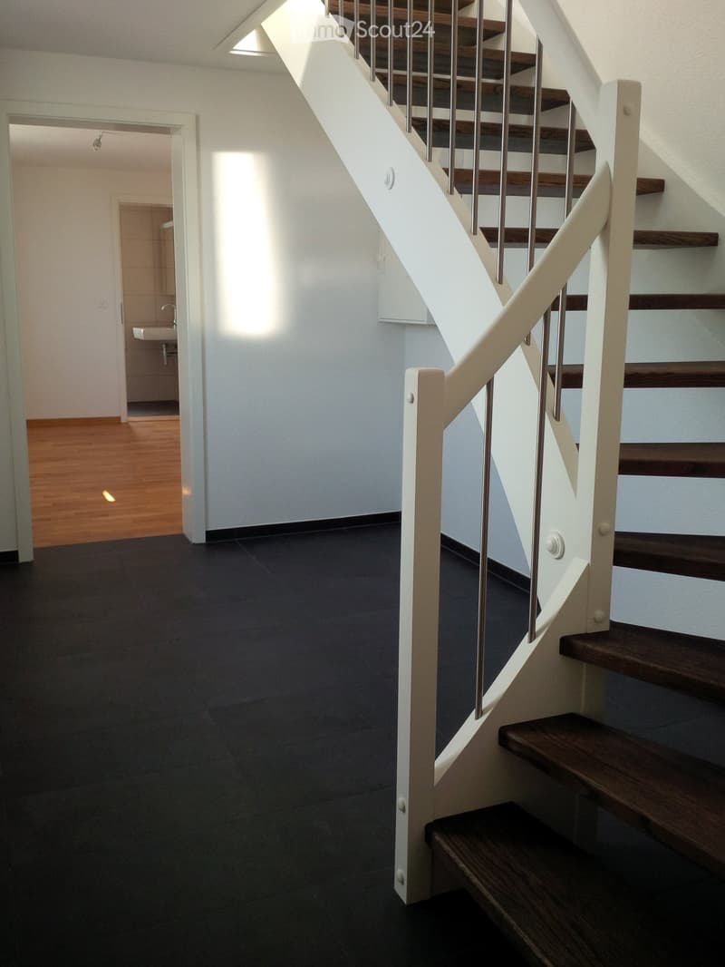 Modern hallway with dark tiled floor, white walls, wooden staircase with metal railing, and a small bathroom visible in the background.