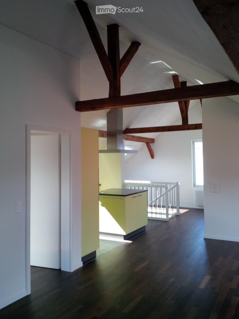 An attic space with exposed wooden beams, hardwood flooring, and a small kitchen area with cabinets and a countertop. The room is empty, with no furniture or personal items visible.