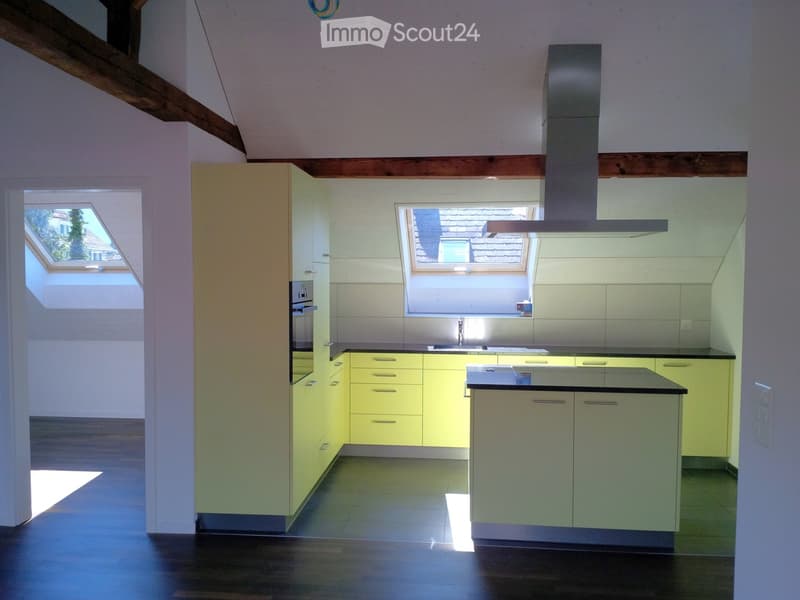Bright kitchen with light yellow cabinets, dark countertops, and a central island. Features exposed wooden beams, skylights, and a modern range hood.