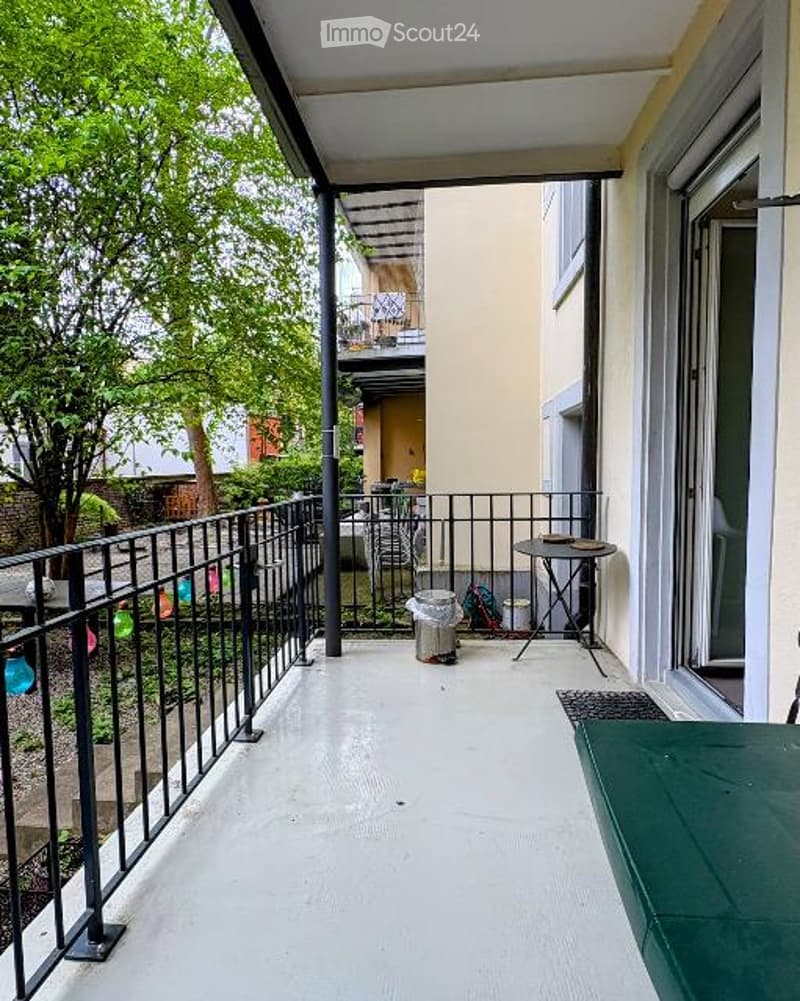 Balcony with white floor, black metal railing, small table, and chair. Green outdoor furniture is partially visible.