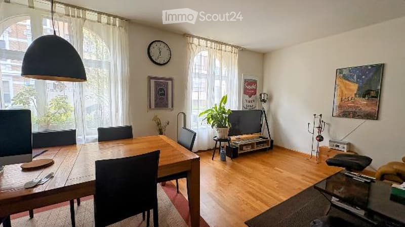 Bright, open-plan living and dining area with wooden flooring, large windows with white curtains, a dining table with chairs, a potted plant, a television, and wall art.