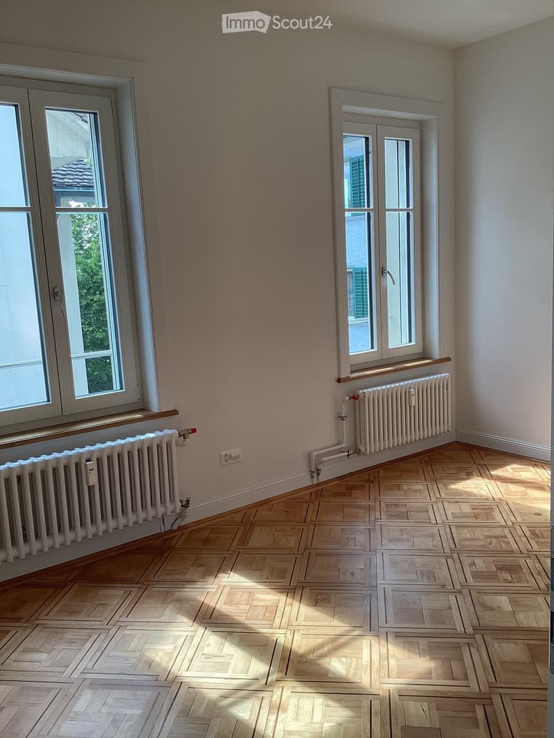 Empty room with wooden parquet flooring, white walls, and two windows with radiators beneath them.