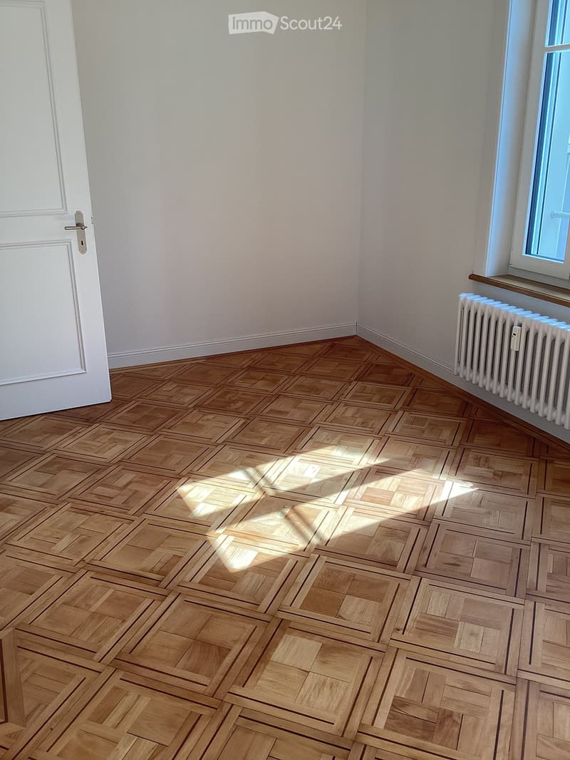 Empty room with wooden parquet flooring, white walls, a white door, and a window with a radiator below it.