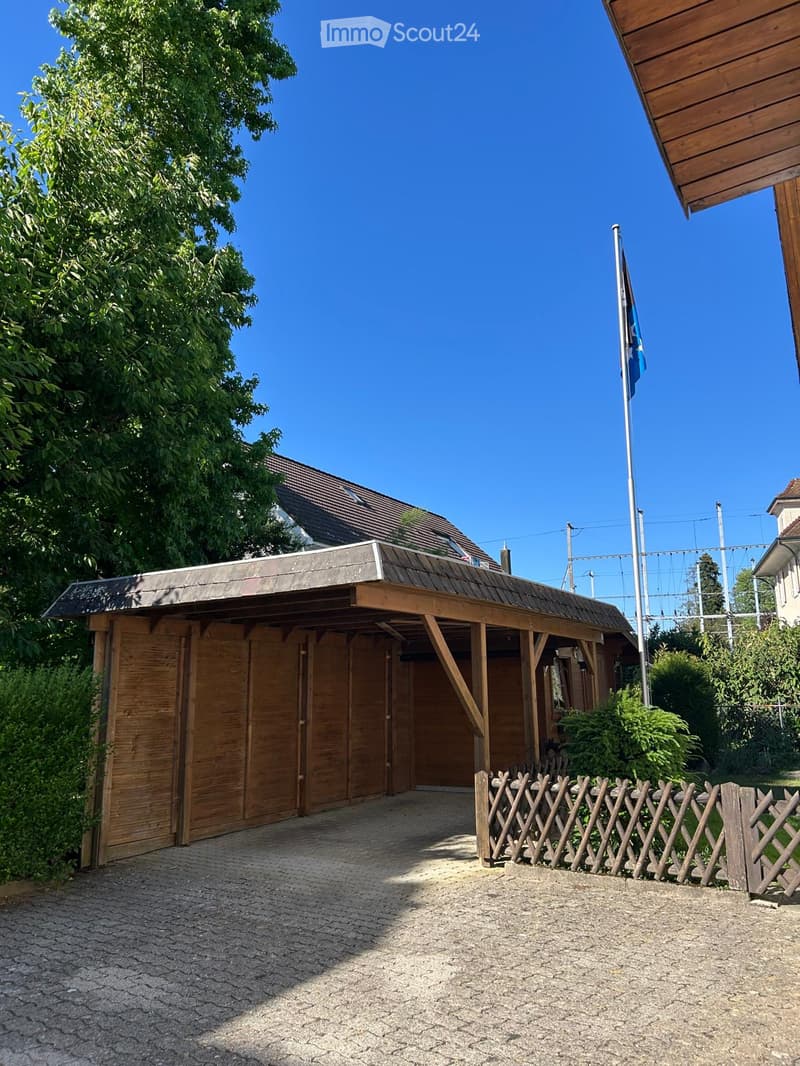 Wooden garage with a slanted roof, surrounded by greenery and a wooden fence.
