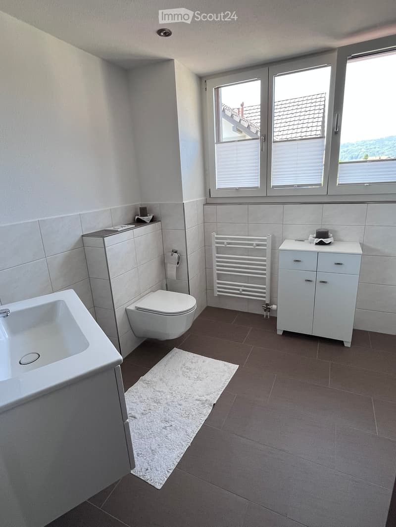 Modern bathroom with a bathtub, toilet, sink, radiator, and a white towel on the floor. The room features large windows with blinds and a view of the roof.