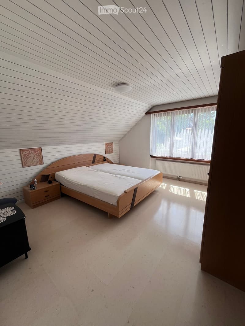 Bedroom with a wooden bed, nightstand, and chair. The room features a slanted ceiling and large window with white curtains.