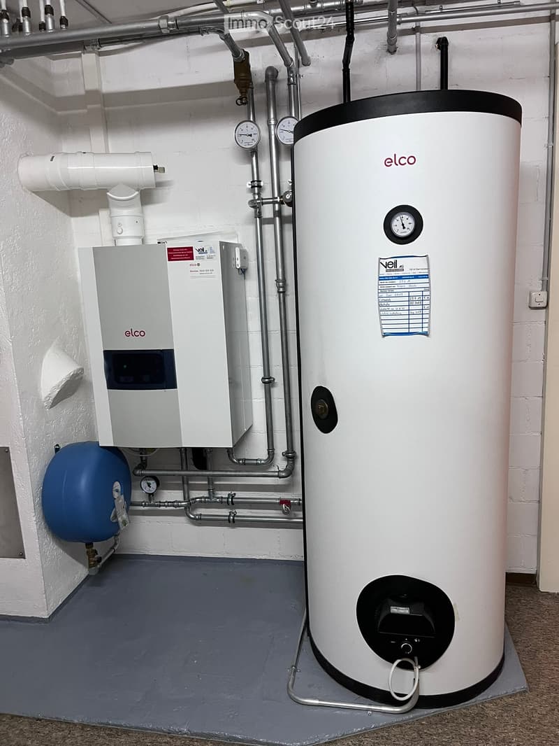 A utility room with a large white water heater, a pressure vessel, control unit, and various pipes and gauges.