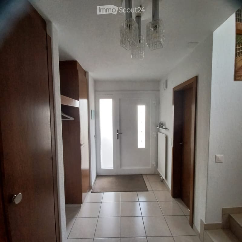 A narrow hallway with tiled flooring, white walls, and wooden doors on both sides. A chandelier hangs from the ceiling, and a small rug is placed in the center of the hallway.