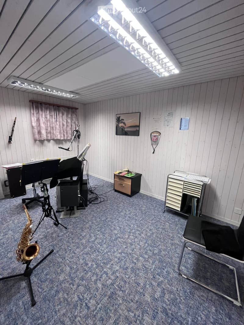 Music room with carpeted floor, slanted ceiling, and various musical instruments including a saxophone, piano, and microphone. There is a chair, a small table, and a cabinet with drawers.