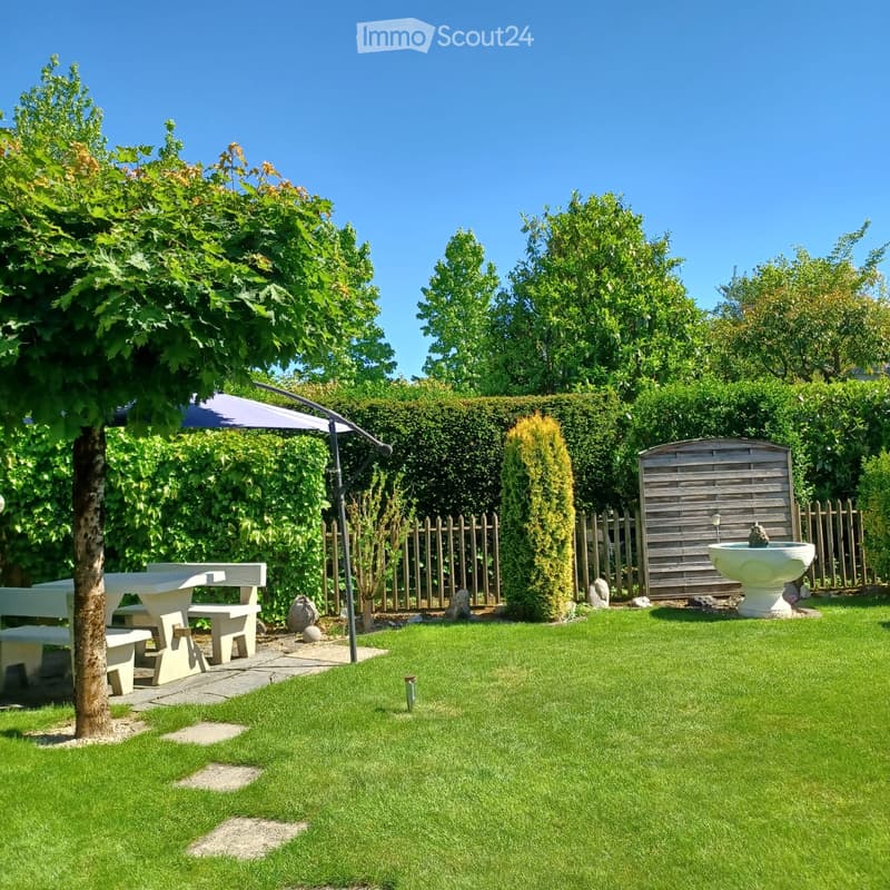 Well-maintained garden with a white picnic table, benches, a fountain, and a large umbrella. The area is enclosed by a wooden fence and surrounded by lush greenery and trees.