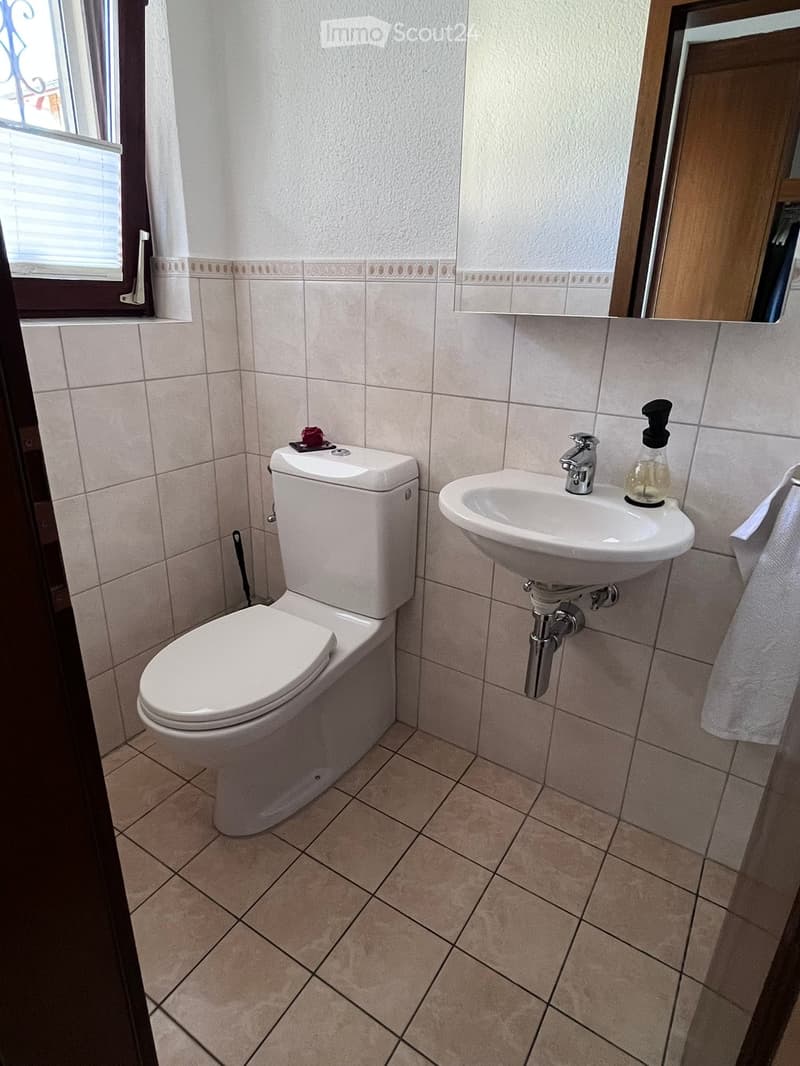 Clean bathroom with white tiled walls and floor, featuring a toilet, sink, and a towel on the side.