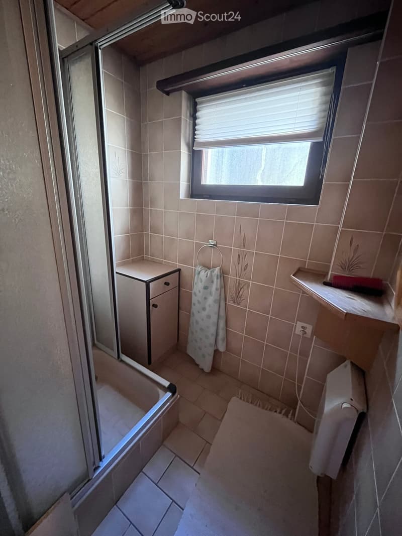 Small bathroom with tiled walls and floor, featuring a bathtub, a small cabinet, a towel, and a window with blinds.
