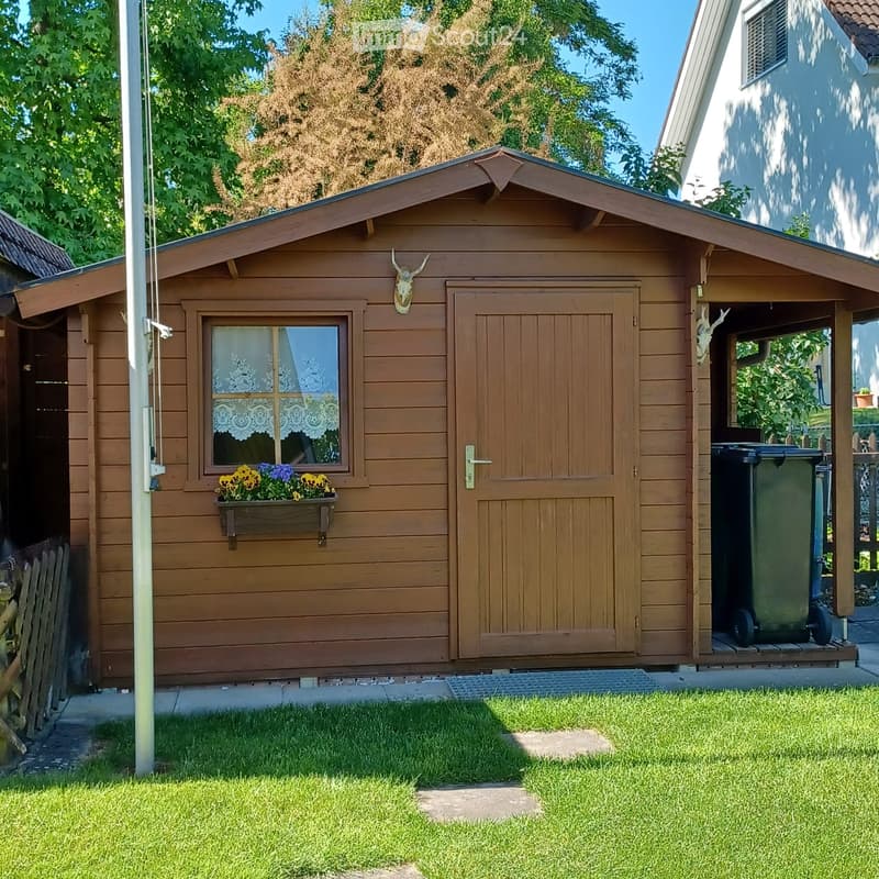 A wooden shed with a pitched roof, a window with lace curtains, a flower box with yellow and purple flowers, and a mounted deer head on the wall. The shed has a closed wooden door and a small open area with a green trash bin.
