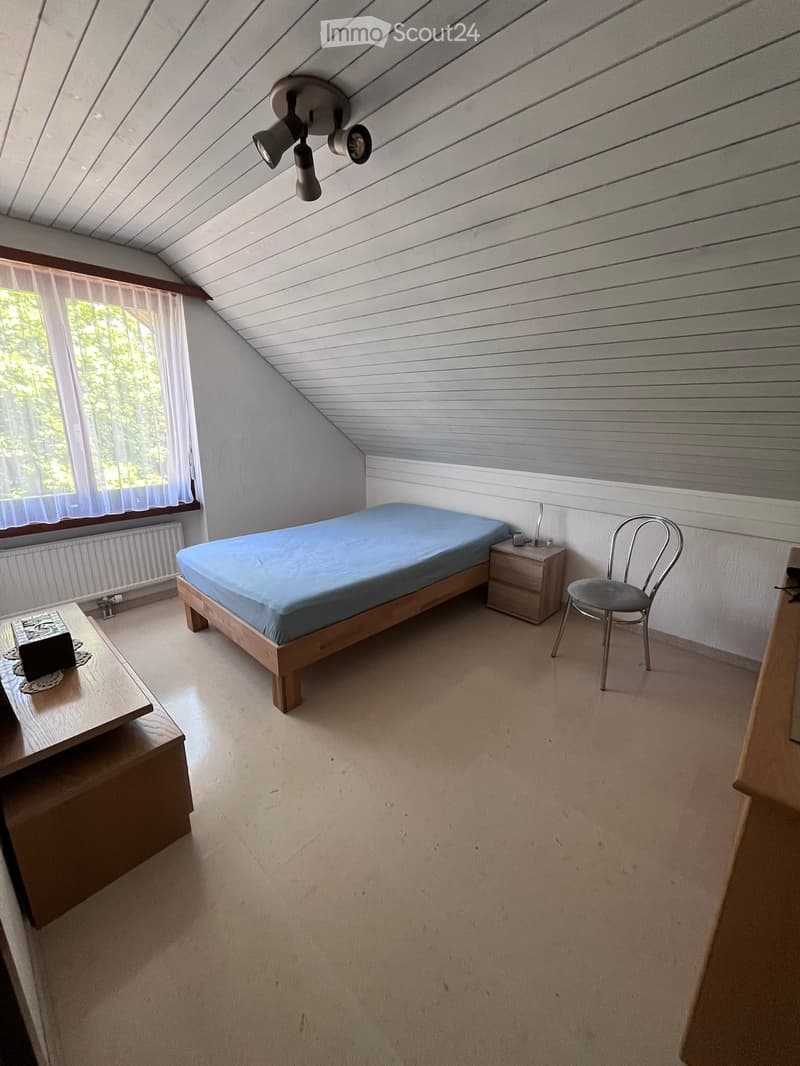 Cozy attic bedroom with slanted wooden ceiling, bed, nightstand, chair, and small table near window with sheer curtains.