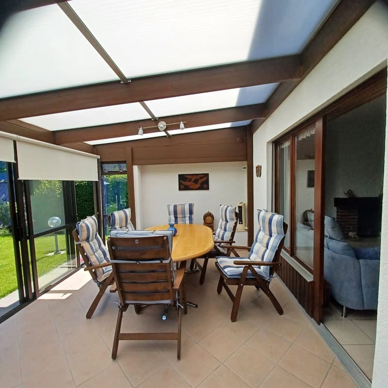 A spacious, covered balcony with a wooden table and six striped chairs, large windows, and a view of a green garden.