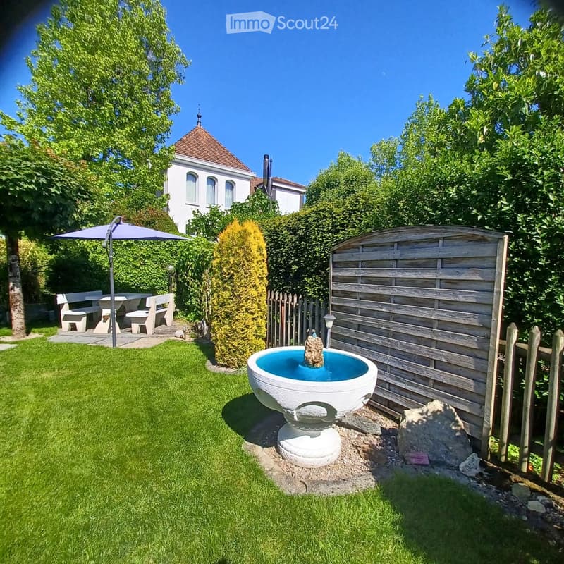Well-maintained garden with a white fountain, a wooden fence, a patio area with a table and chairs, and a large umbrella. The garden is surrounded by lush greenery and trees, with a house visible in the background.