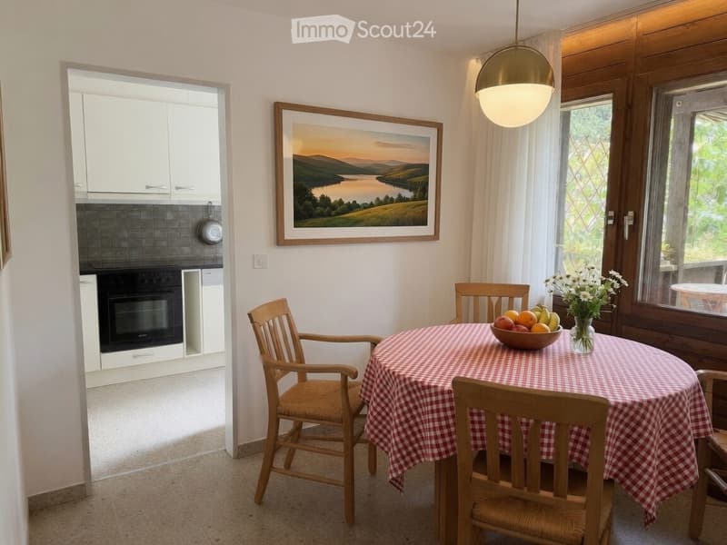 Bright kitchen with a dining area, featuring a round wooden table with a checkered tablecloth, four wooden chairs, a bowl of fruit, a vase with white flowers, and a framed landscape painting on the wall. The kitchen area includes white cabinets, a black-t