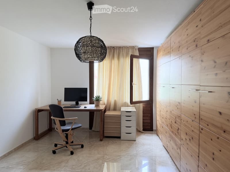 A modern, well-lit study or home office with a wooden desk, ergonomic chair, computer setup, and a large wooden wardrobe. The room features a hanging spherical lamp, a window with curtains, and a small potted plant on the desk.