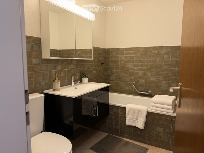 Modern bathroom with dark green tiles, white sink, black cabinet, bathtub, and neatly stacked towels.