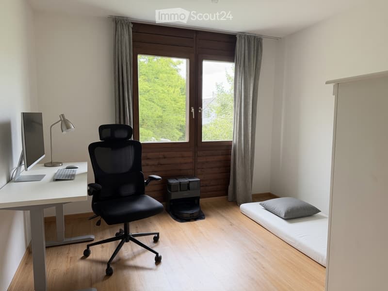 A modern, well-lit room with a desk setup, a comfortable chair, a small bed, and a robot vacuum. The room features large windows with curtains and wooden flooring.