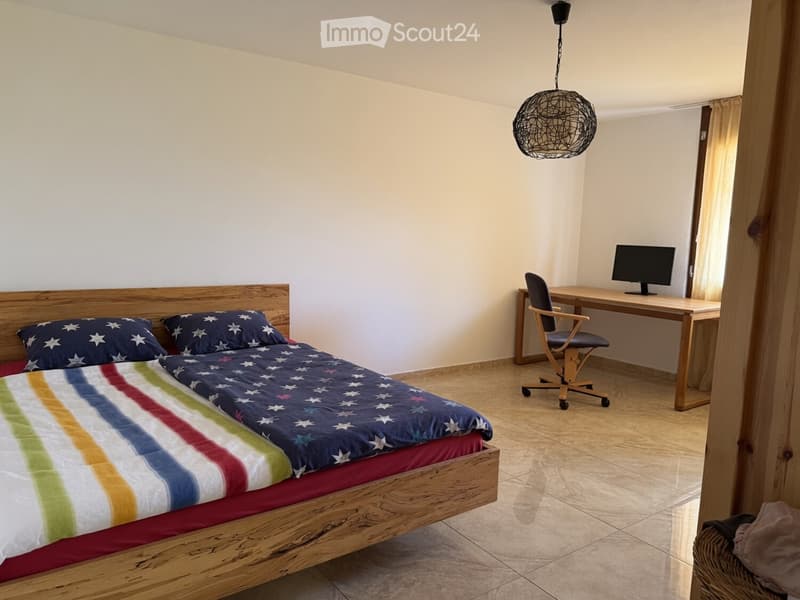 Cozy bedroom with a wooden bed, colorful striped bedding, a small desk with a computer, and a hanging woven light fixture.