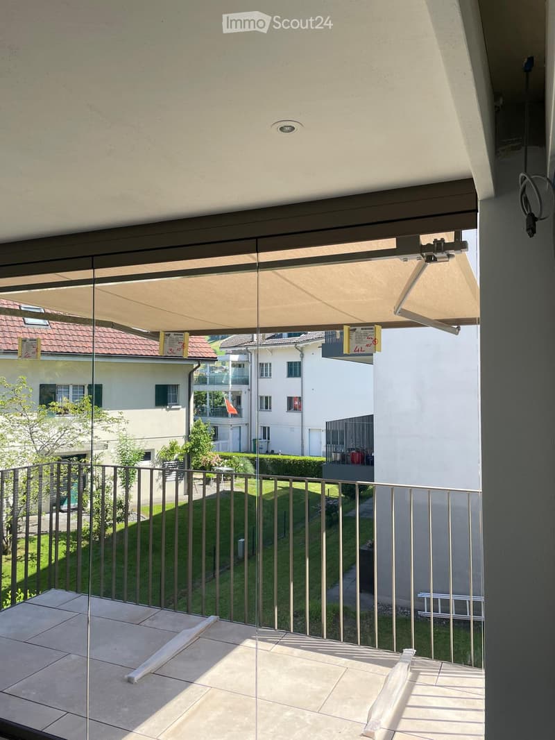 Balcony with tiled floor, metal railing, and retractable awning, overlooking a green lawn and neighboring houses.