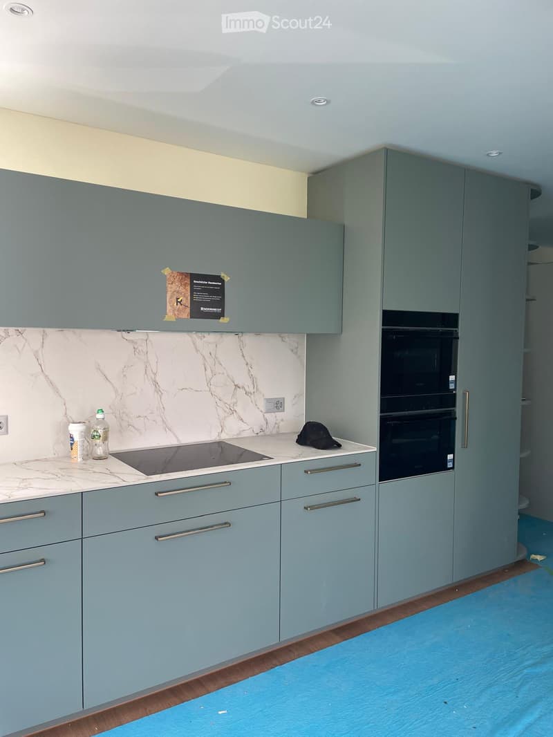 Modern kitchen with light blue cabinets, marble backsplash, induction stove, and double oven. Countertop has a few items including a bottle and a hat.