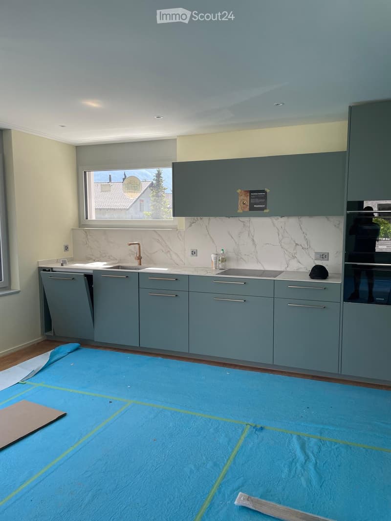 Modern kitchen with grey cabinets, marble backsplash, and stainless steel sink. The floor is covered with a blue protective sheet.