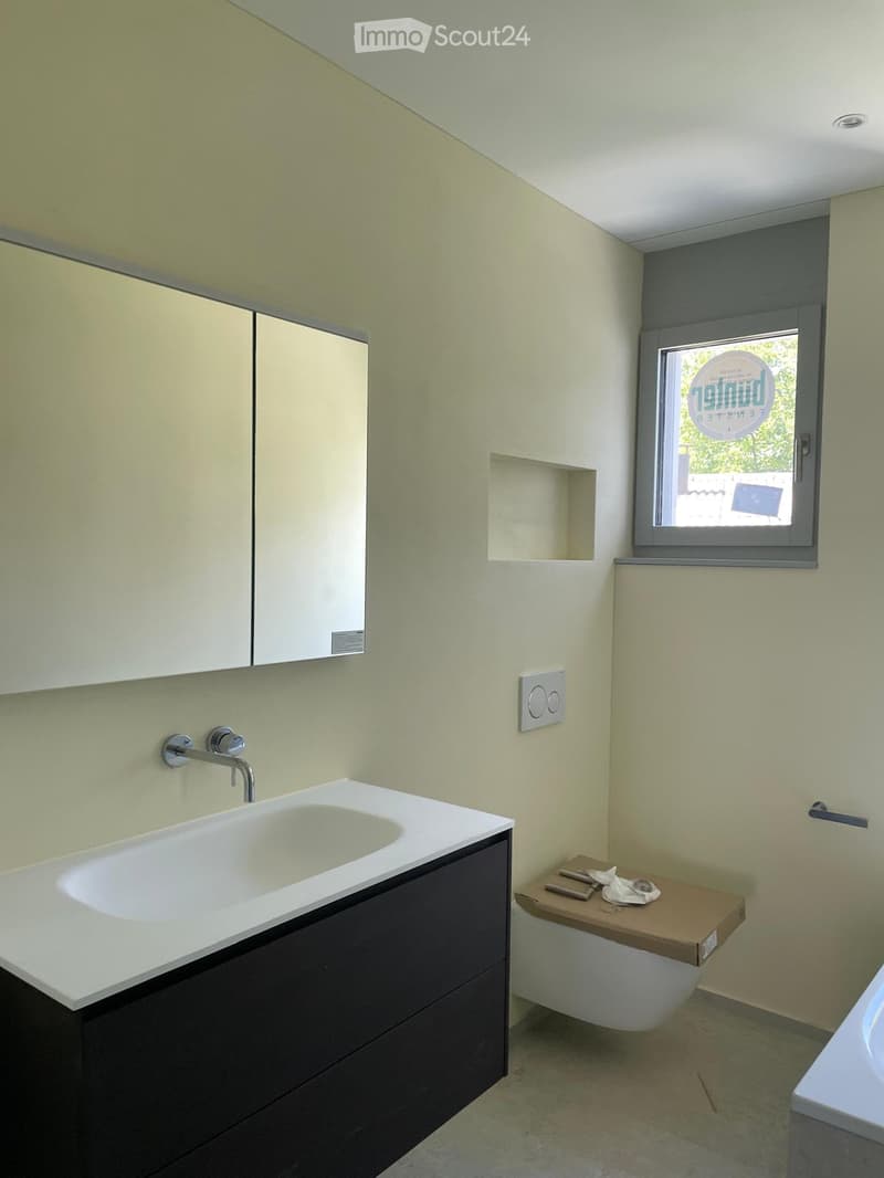 Modern bathroom with a white sink, dark cabinet, mirror, and a toilet with a wooden seat cover. The walls are light-colored, and there is a window with a view of greenery.