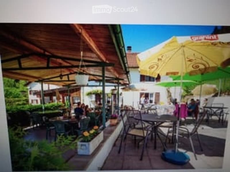 Outdoor seating area with tables and chairs under umbrellas, surrounded by greenery and a clear blue sky.