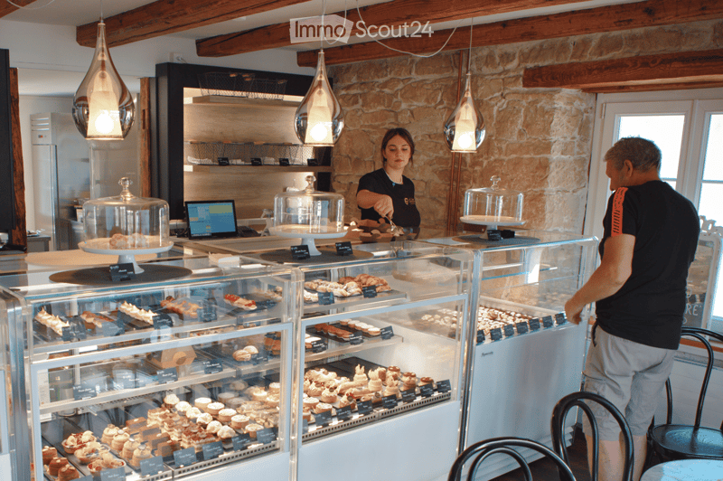 Bakery interior with exposed wooden beams, stone walls, and glass display cases filled with pastries and cakes. A staff member is serving a customer.