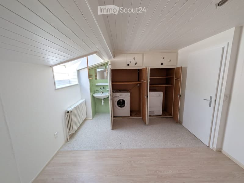 A small, well-lit room with a washing machine, sink, radiator, and built-in cabinets. The space features light wooden flooring and white walls.