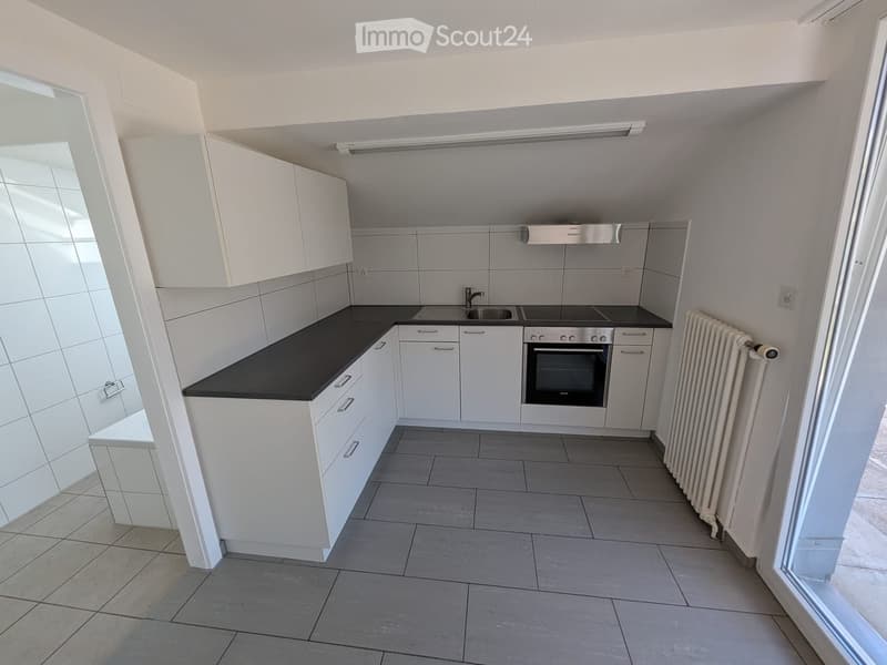 Modern kitchen with white cabinets, black countertops, tiled walls, and a built-in oven. Features a sink, radiator, and large windows allowing natural light.