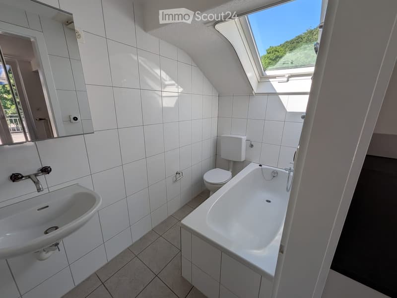 Modern bathroom with white tiles, a sink, bathtub, toilet, and a large mirror. Features a skylight window offering a view of greenery.