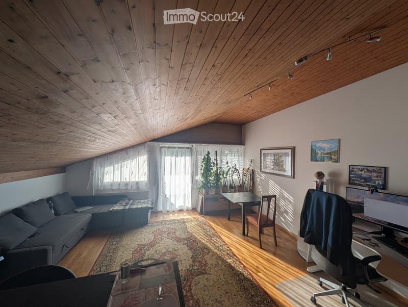 A cozy attic room with wooden beams, a sofa, a desk with a laptop, a chair, and a large window with curtains. The room features a patterned rug and several potted plants.