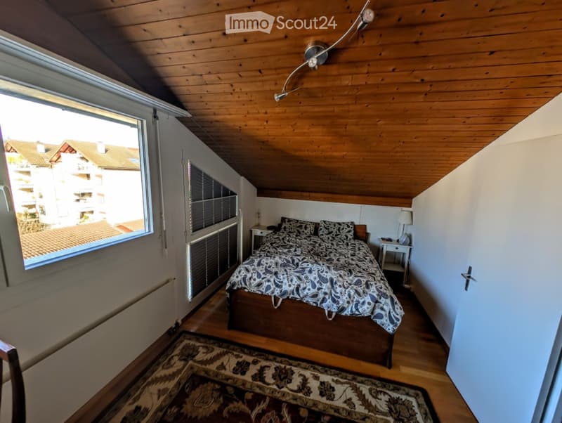 Cozy attic bedroom with wooden ceiling, large window offering a view, bed with patterned bedding, nightstand, and a patterned rug.