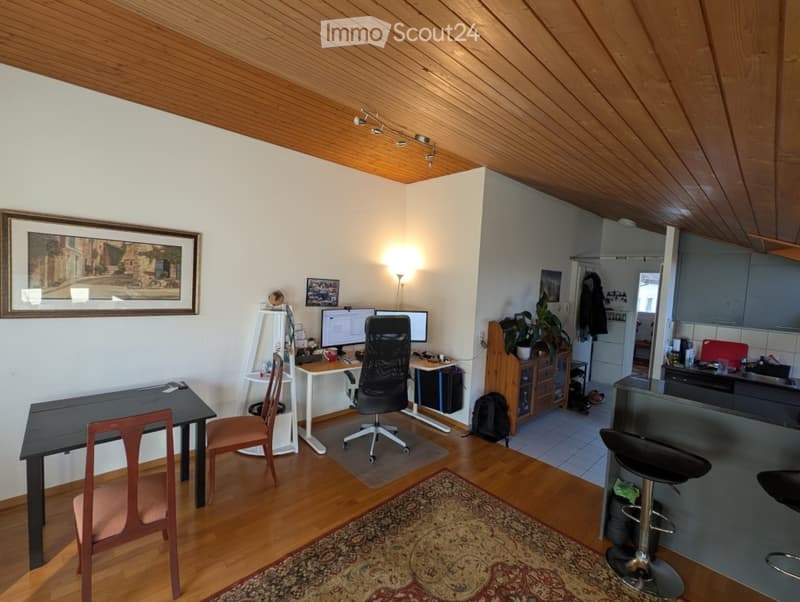 A modern, well-lit room with wooden flooring and a slanted wooden ceiling. It features a desk with a computer, a comfortable chair, a small table, a stool, and a large picture frame on the wall. A small kitchen area is visible in the background with a cou