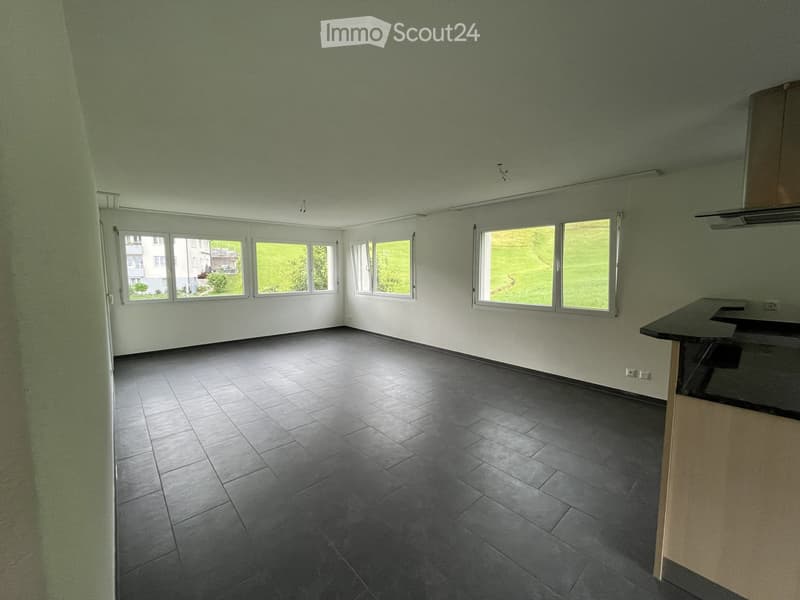Empty room with grey tiled floor, white walls, and large windows offering a view of greenery. A small kitchen counter is visible on the right side.
