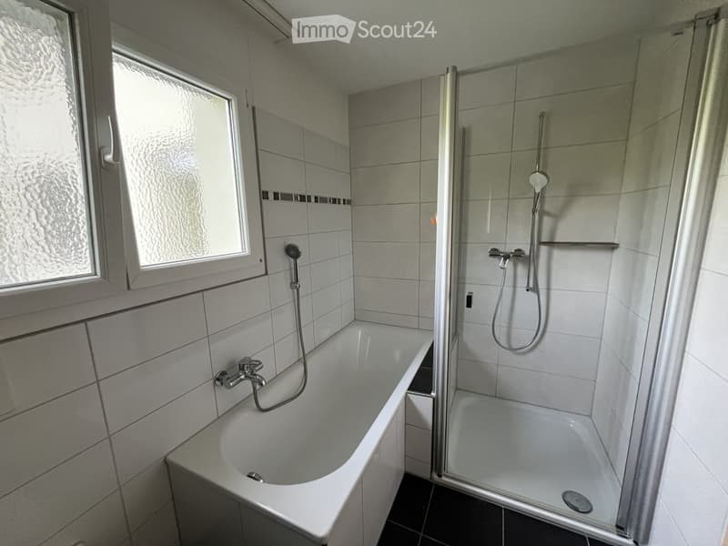 Modern bathroom with a bathtub, shower, and large window. Features white tiles, a glass shower enclosure, and a modern faucet.